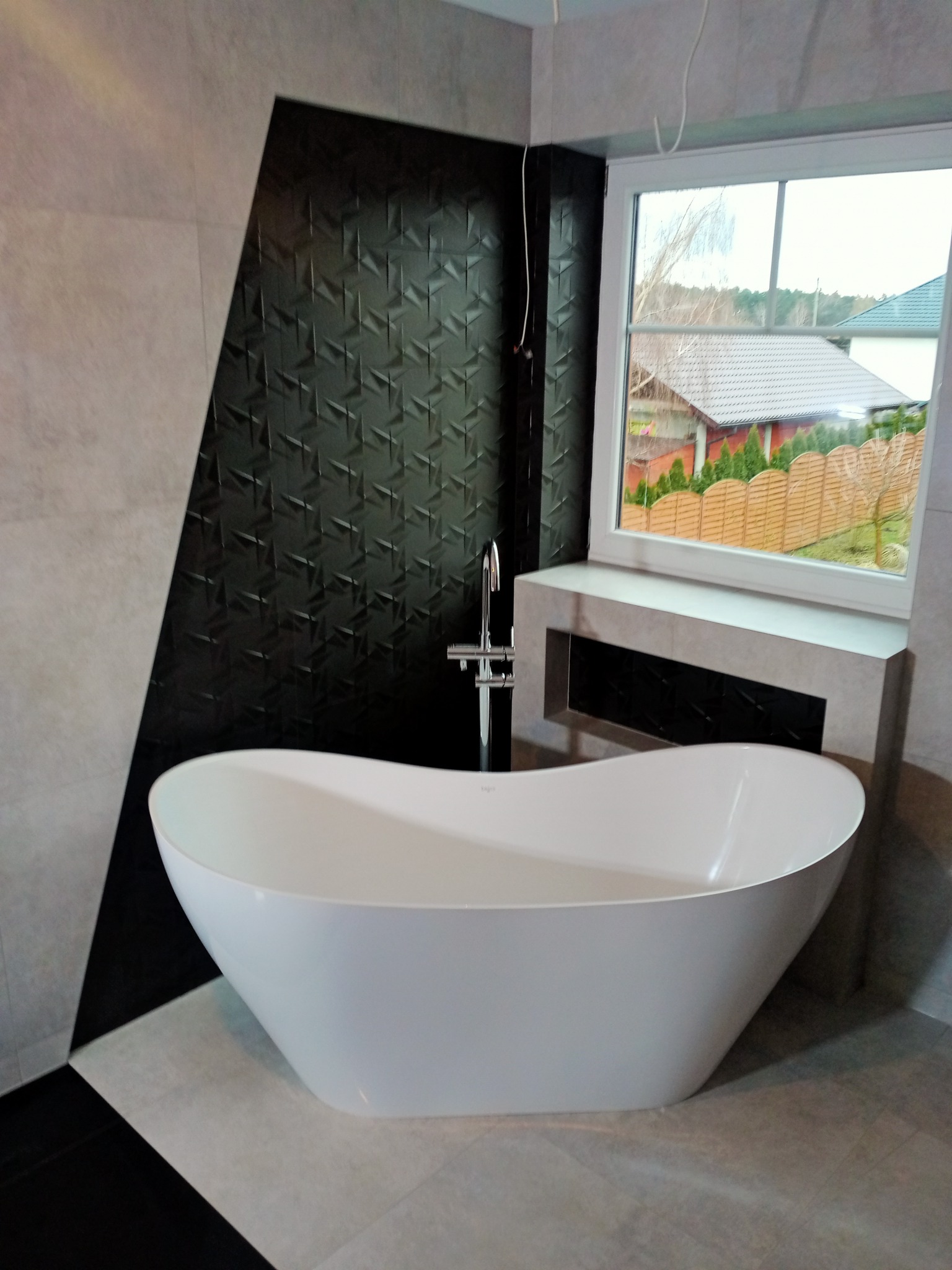 Nowoczesna łazienka z wolnostojącą białą wanną na tle ściany z czarną, geometryczną mozaiką i dużym oknem z widokiem na ogród.