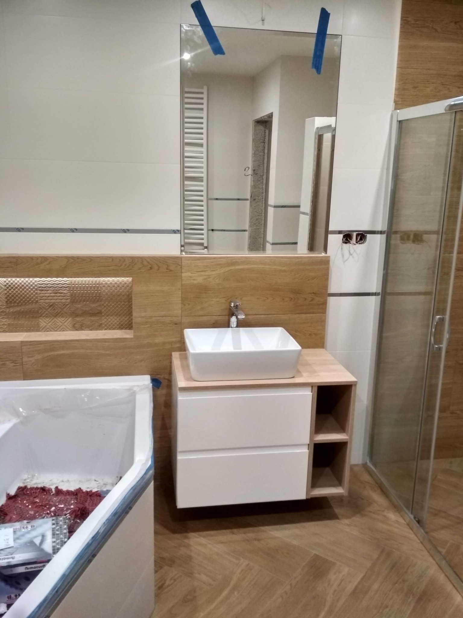 Nowoczesna łazienka w trakcie wykończenia: biała umywalka nablatowa na szafce z szufladami, kabina prysznicowa ze szkła, lustro z niebieską taśmą zabezpieczającą, drewnopodobne płytki na ścianach...