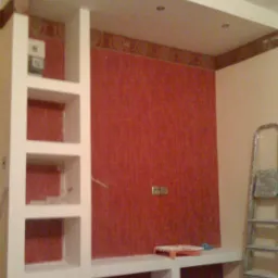 Wnęka z półkami i podwieszanym sufitem w pokoju z czerwoną tapetą, widoczna drabina malarska i narzędzia malarskie.