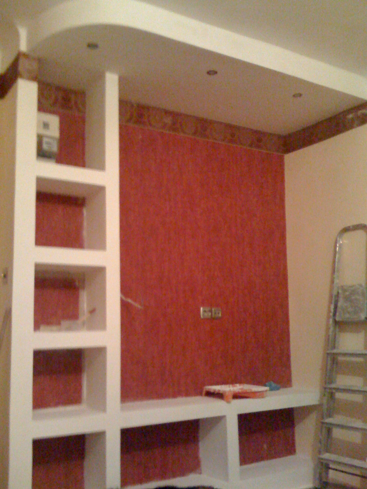 Wnęka z półkami i podwieszanym sufitem w pokoju z czerwoną tapetą, widoczna drabina malarska i narzędzia malarskie.