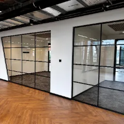 Przeszklone ścianki działowe z czarnymi ramami w nowoczesnym biurze, widok na sąsiednie pomieszczenia z wykładziną dywanową, parkiet w jodełkę na pierwszym planie, instalacje pod sufitem.