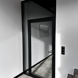 HoldCraft Sp. z o. o. - Szklane drzwi wewnętrzne z czarną ramą w minimalistycznym wnętrzu, widoczny fragment sofy i włączniki na ścianie.