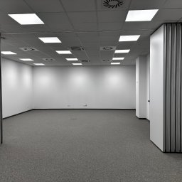 HoldCraft Sp. z o. o. - Puste, jasne biuro z szarą wykładziną i składaną ścianą działową, widoczne gniazdka elektryczne przy podłodze.