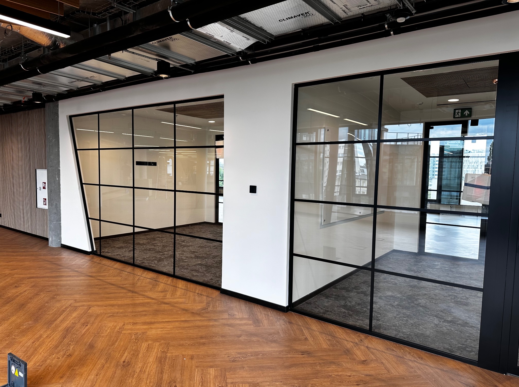 Przeszklone ścianki działowe z czarnymi ramami w nowoczesnym biurze, widok na sąsiednie pomieszczenia z wykładziną dywanową, parkiet w jodełkę na pierwszym planie, instalacje pod sufitem.