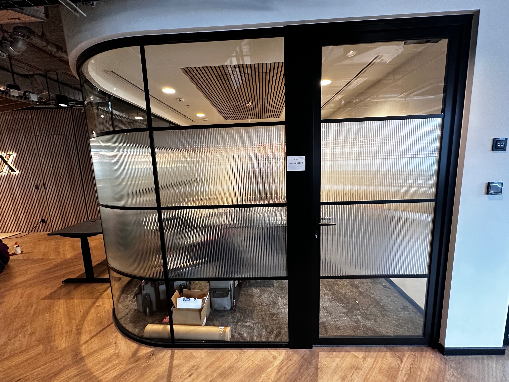 Nowoczesna szklana ścianka działowa z czarnymi ramami i ryflowanym szkłem w biurze, widoczne kartony i torby wewnątrz.