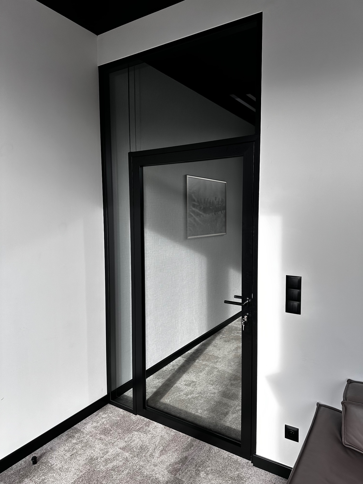 Szklane drzwi wewnętrzne z czarną ramą w minimalistycznym wnętrzu, widoczny fragment sofy i włączniki na ścianie.