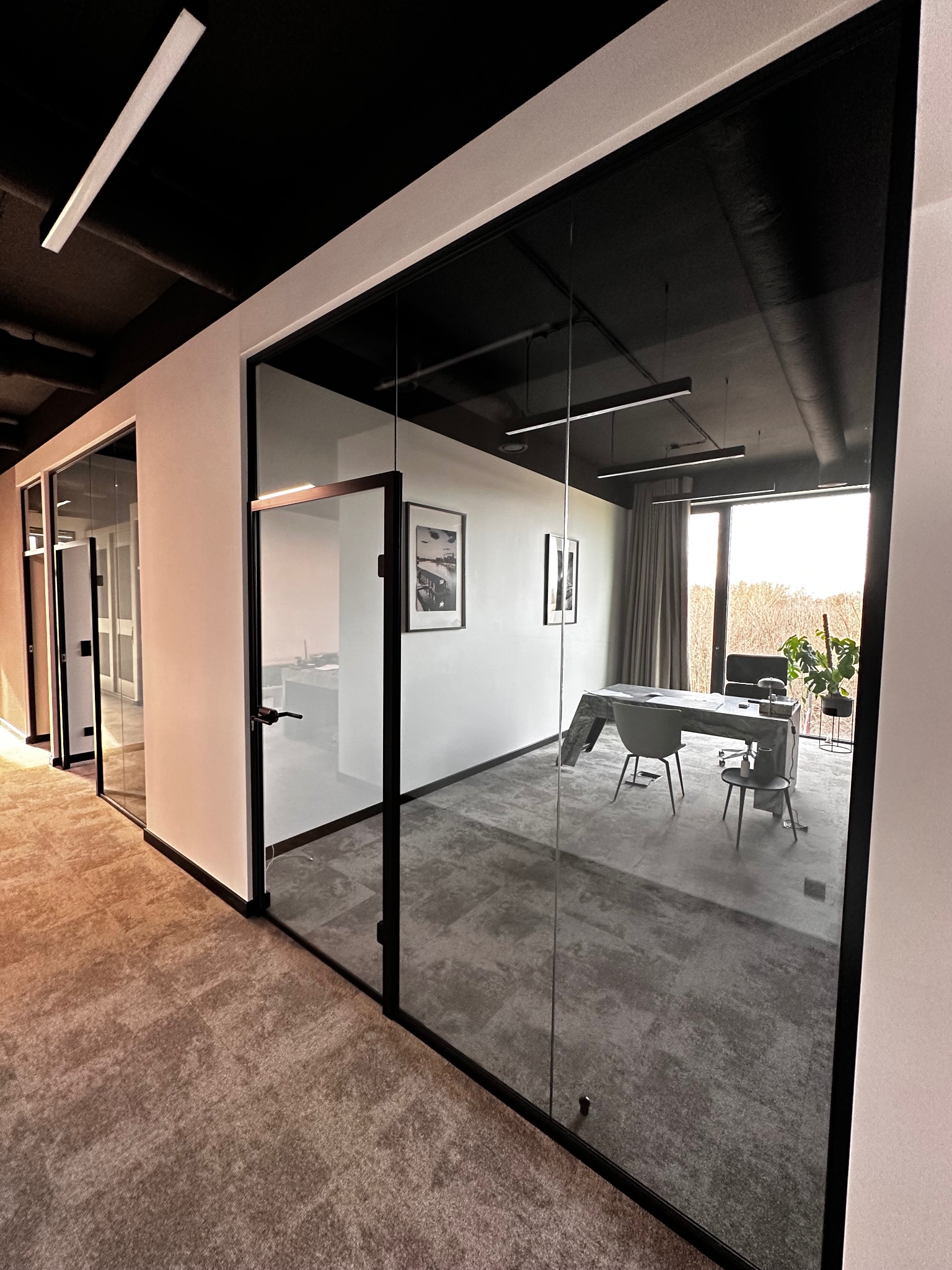 Nowoczesne biuro z przeszklonymi ścianami i drzwiami w czarnych ramach, widok na przestrzeń biurową z designerskim biurkiem i krzesłami, oświetlenie liniowe na suficie.