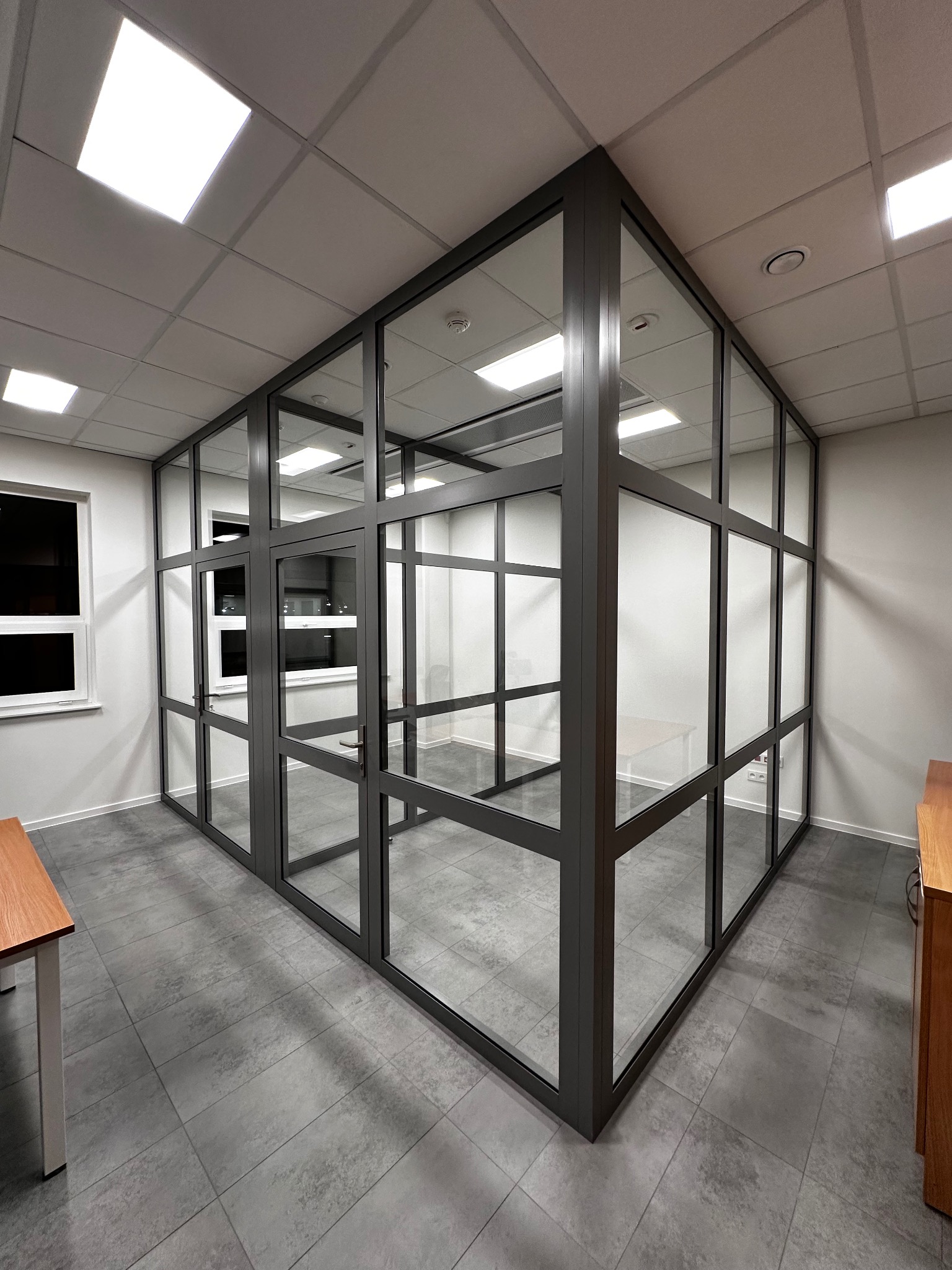 Szklana ścianka działowa z drzwiami w biurze, szara rama, widok z perspektywy, szare płytki podłogowe.