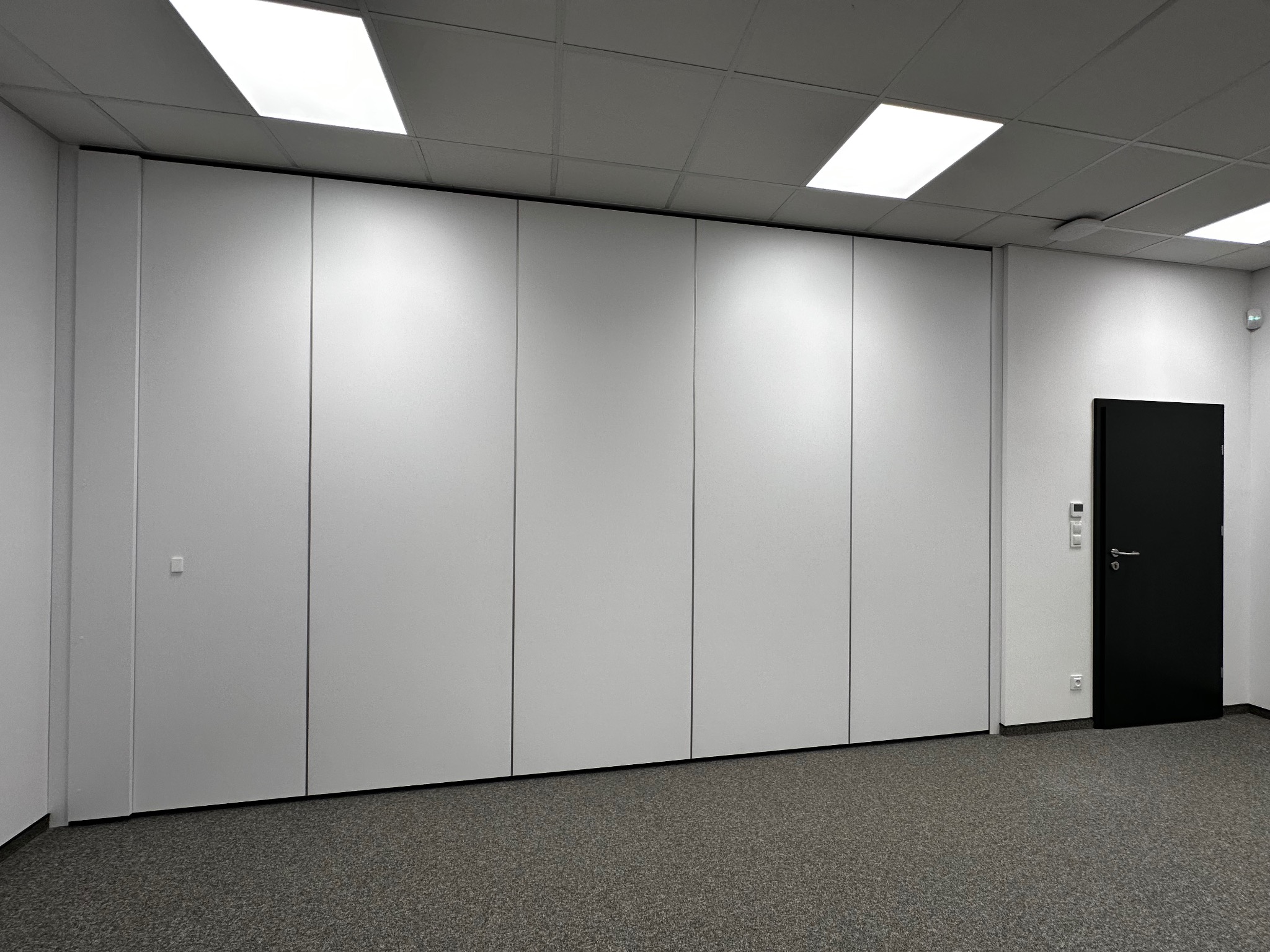 Składana ściana działowa z paneli w biurze, widoczna czarna drzwi wejściowe po prawej stronie i szara wykładzina podłogowa.