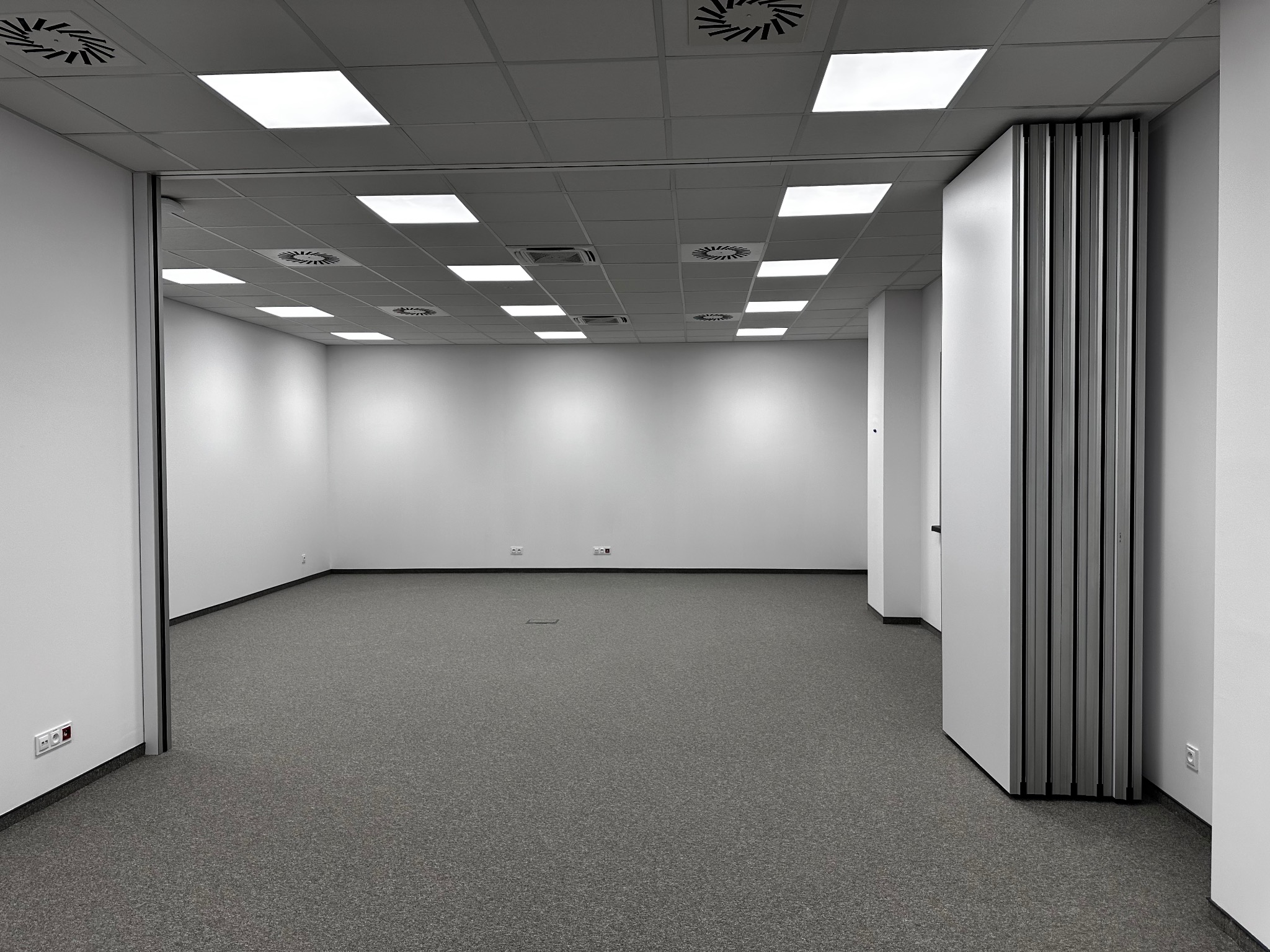 Puste, jasne biuro z szarą wykładziną i składaną ścianą działową, widoczne gniazdka elektryczne przy podłodze.