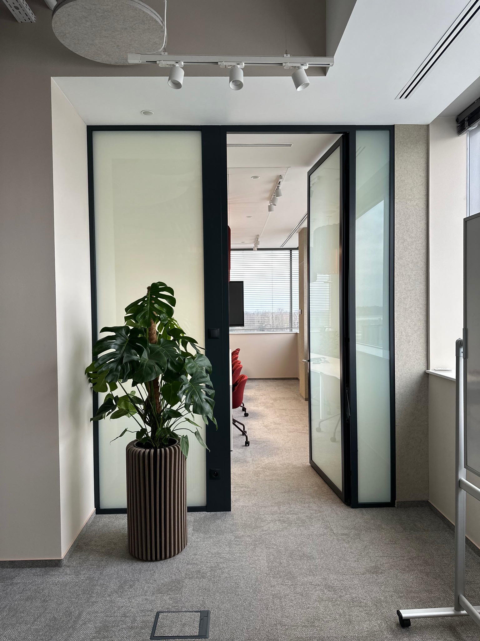 Nowoczesne biuro z przeszklonymi drzwiami przesuwnymi w czarnej ramie, widok na salę konferencyjną z czerwonymi krzesłami, na pierwszym planie roślina Monstera w prążkowanej donicy.