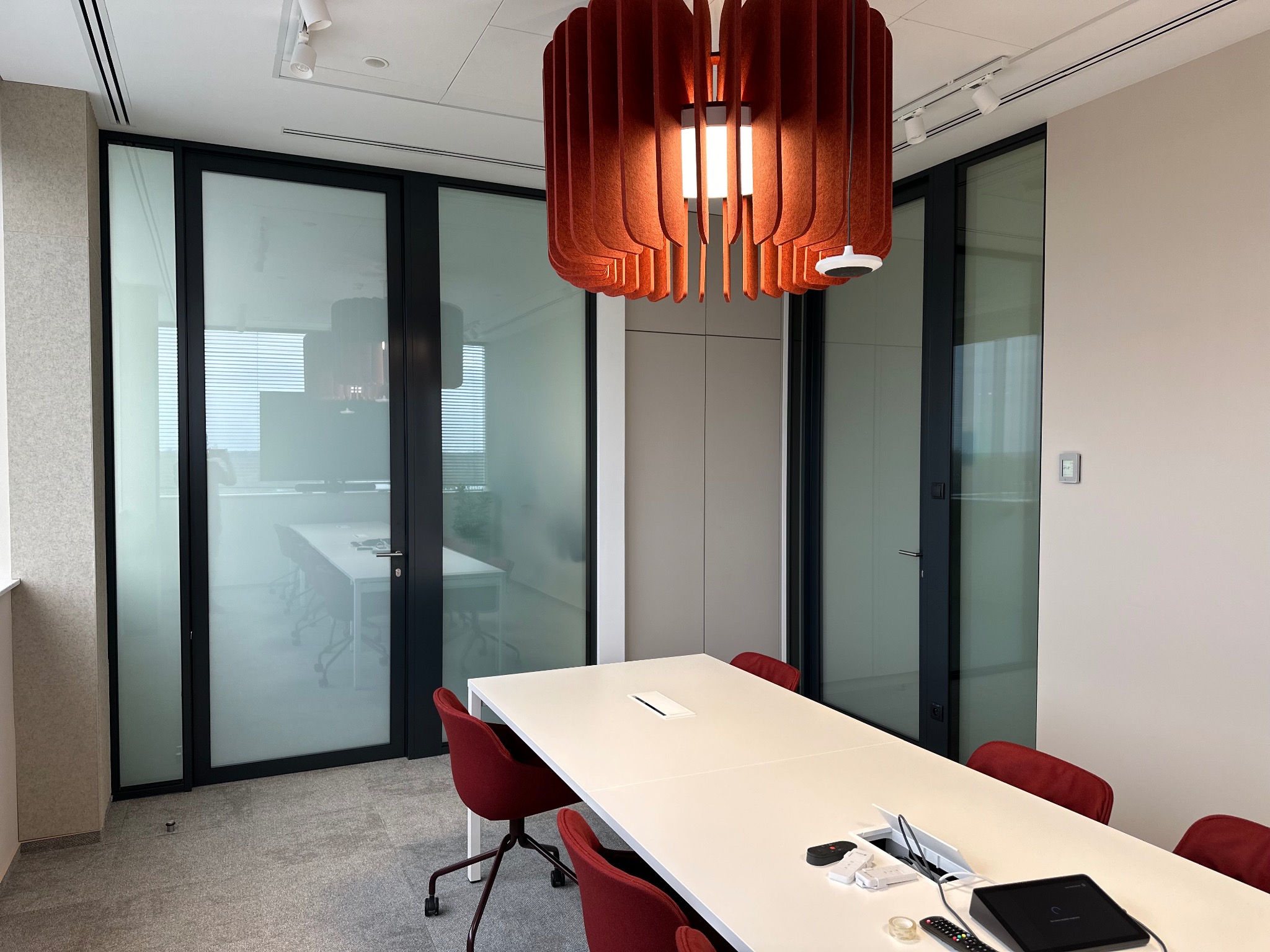 Nowoczesne biuro z przeszklonymi ścianami działowymi, dużym stołem konferencyjnym i oryginalną, pomarańczową lampą wiszącą, widoczne elementy wyposażenia biurowego na stole.