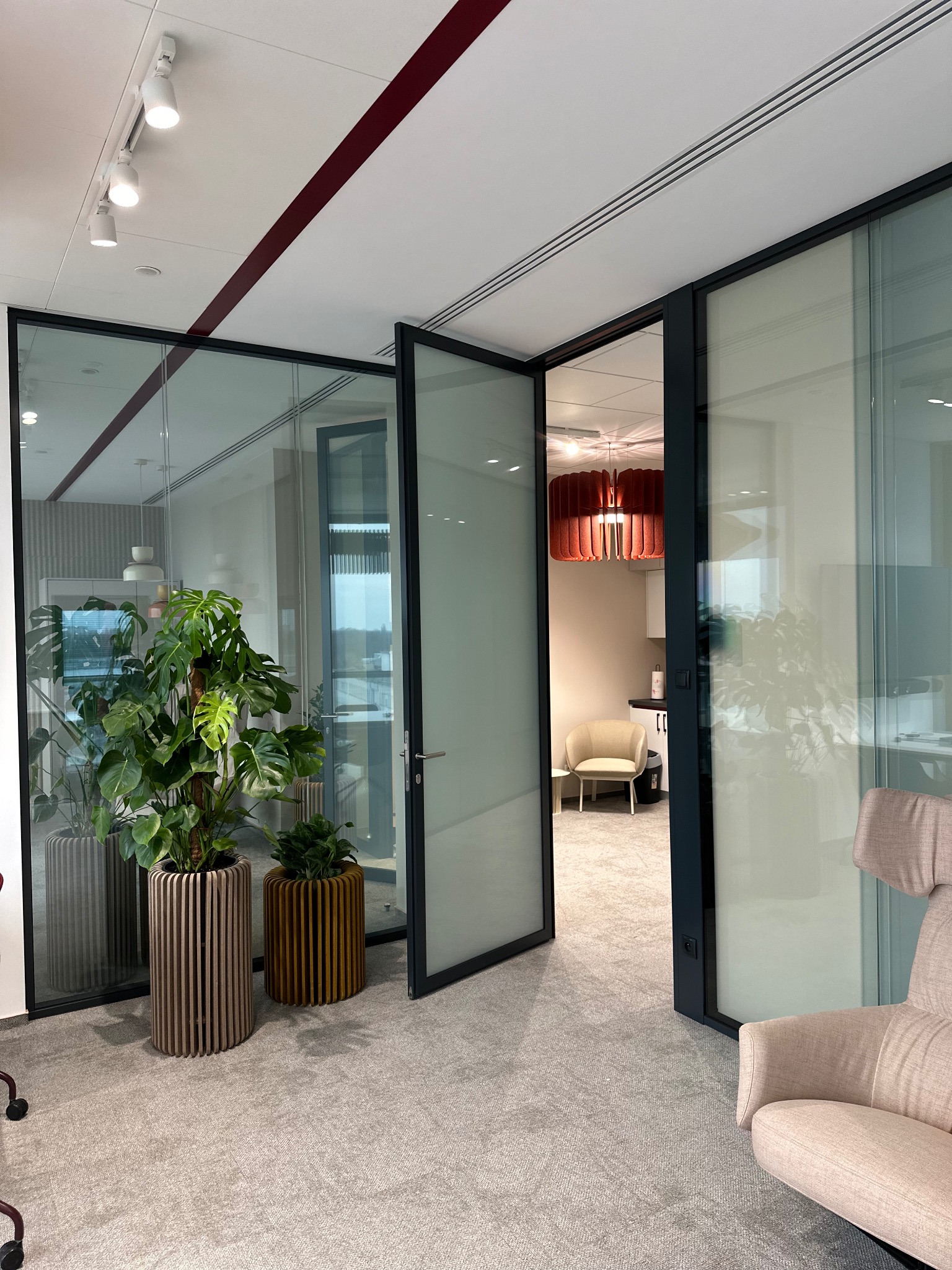 Nowoczesne biuro w Katowicach z przeszklonymi ścianami działowymi, otwartymi drzwiami i roślinami w ozdobnych donicach, widok na wnętrze z designerską lampą i krzesłem.