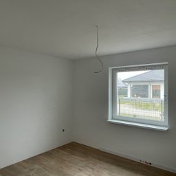 Kon-Stw Kompleksowe wykończenia wnętrz - Pusty pokój z drewnopodobną podłogą, świeżo pomalowanymi białymi ścianami i białym oknem z widokiem na dom i ogród. Z sufitu zwisa kabel.