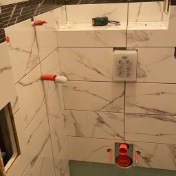 Kon-Stw Kompleksowe wykończenia wnętrz - Łazienka w trakcie remontu: białe płytki marmurowe z czarnym pasem, widoczne rury i instalacja podtynkowa, przymocowany stelaż podtynkowy WC, poziomica oraz resztki zaprawy na półce.