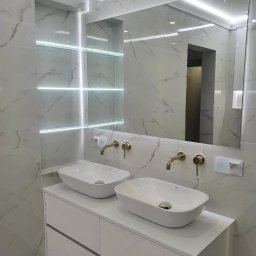 Kon-Stw Kompleksowe wykończenia wnętrz - Nowoczesna łazienka z dwoma umywalkami nablatowymi, złotą armaturą i podświetlanym lustrem w marmurowej okładzinie.