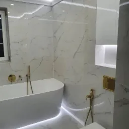 Nowoczesna łazienka z białymi marmurowymi płytkami, podświetlaną wanną wolnostojącą i złotą armaturą, oświetlenie LED pod wanną i w półce ściennej.