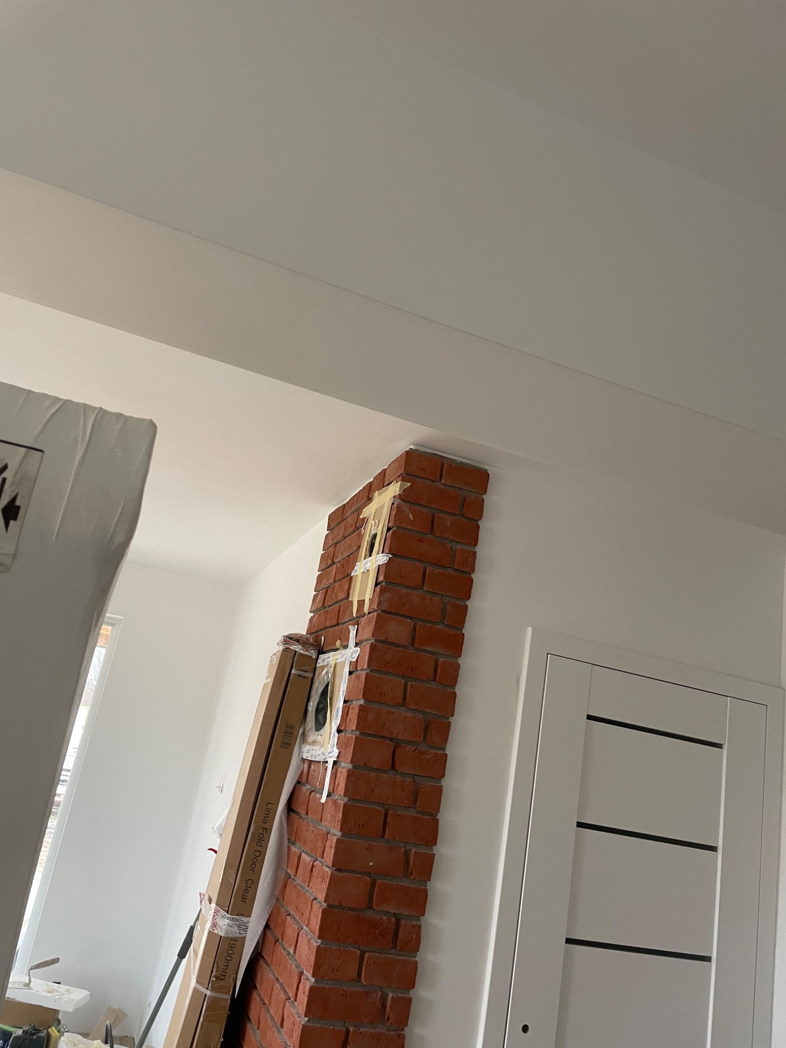 Ceglany filar z widocznymi śladami taśmy malarskiej, przylegający do białej ściany i sufitu, obok zapakowane drzwi opierające się o ścianę, w tle białe, świeżo pomalowane wnętrze.