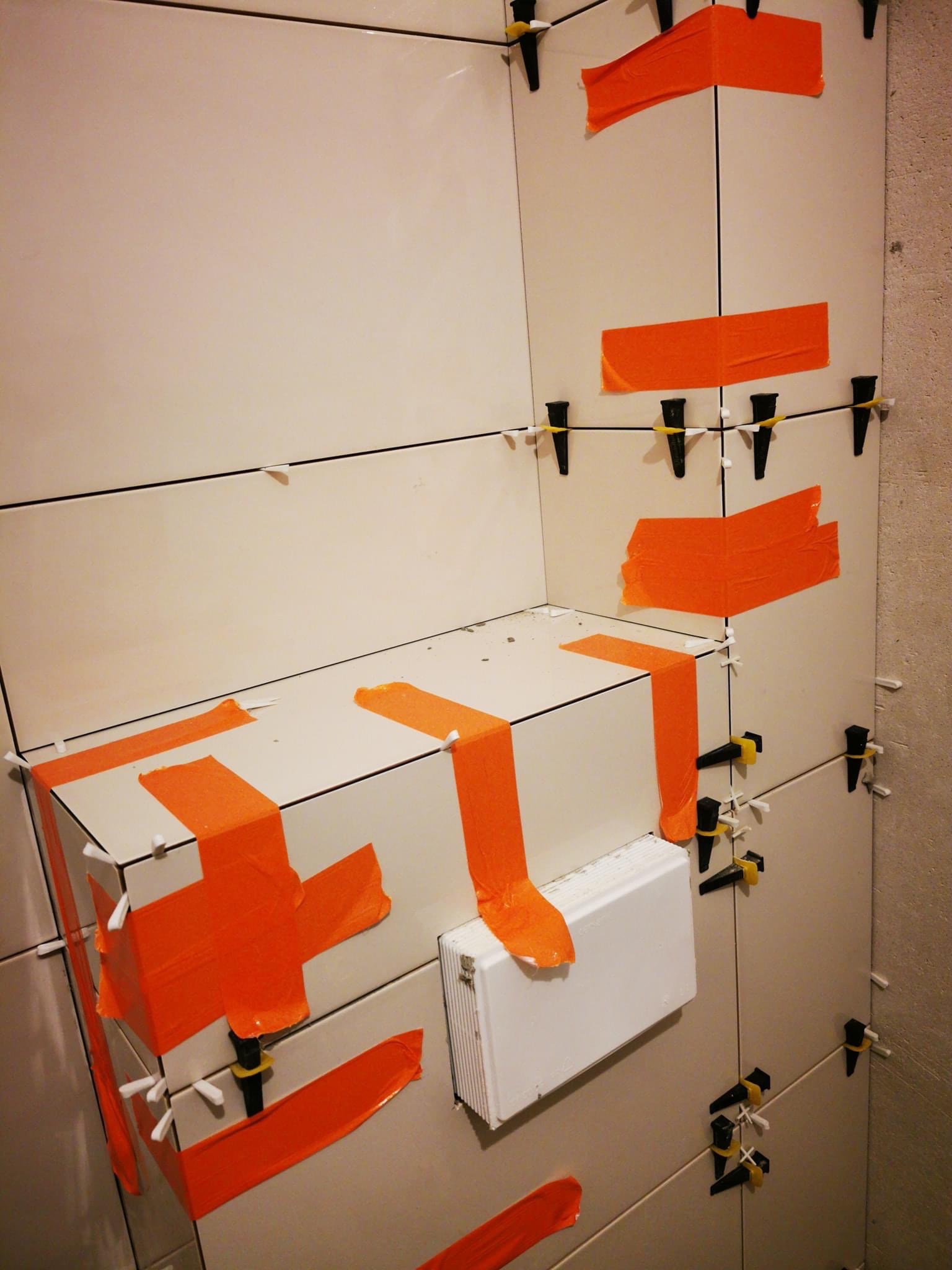 Białe płytki ścienne w trakcie montażu, zabezpieczone pomarańczową taśmą, z widocznymi elementami poziomującymi i fragmentem zabudowy.