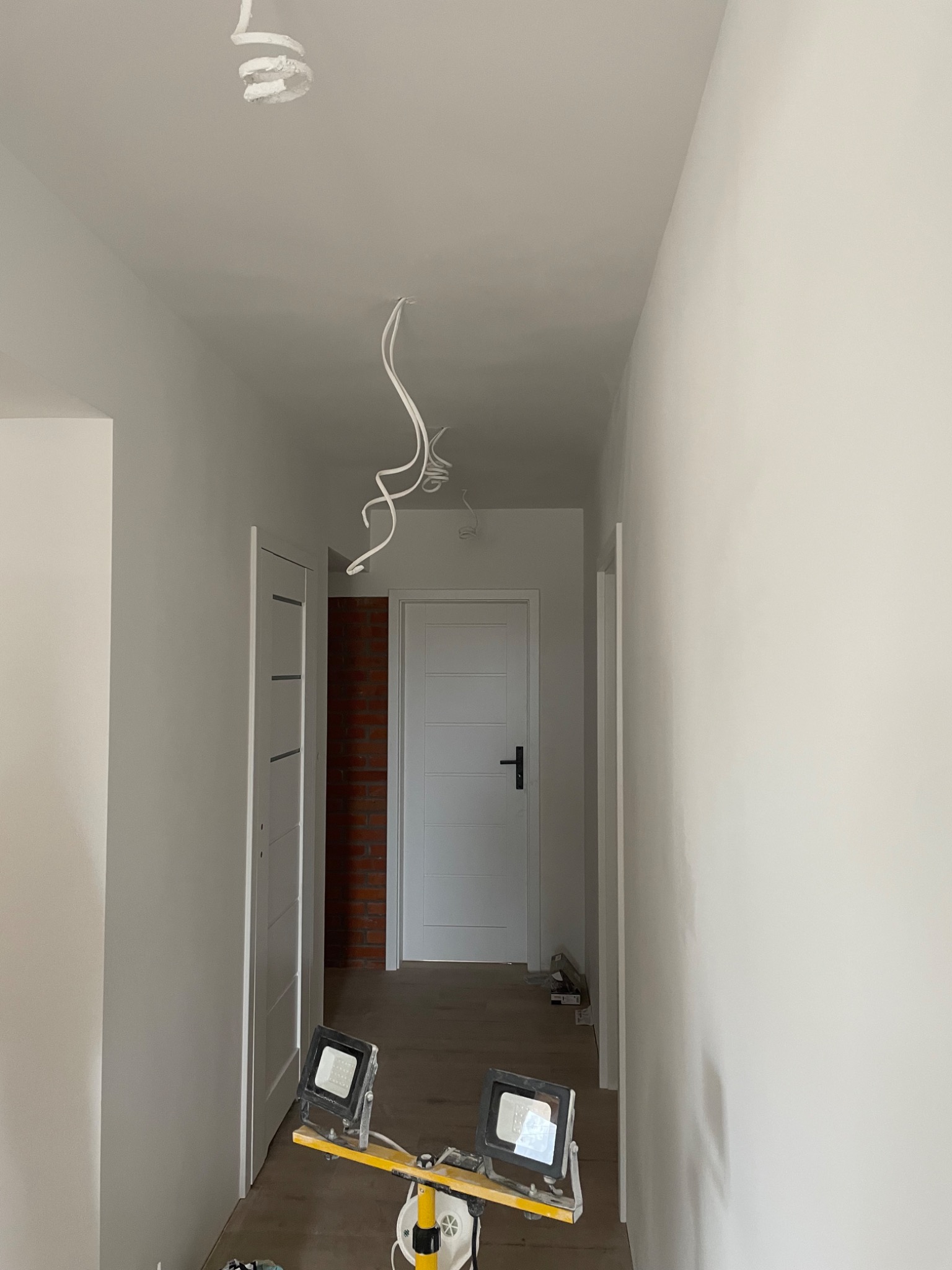 Korytarz w trakcie odświeżania ścian na biało, widoczne wiszące przewody, reflektory halogenowe na podłodze i fragment ściany z cegły w tle.