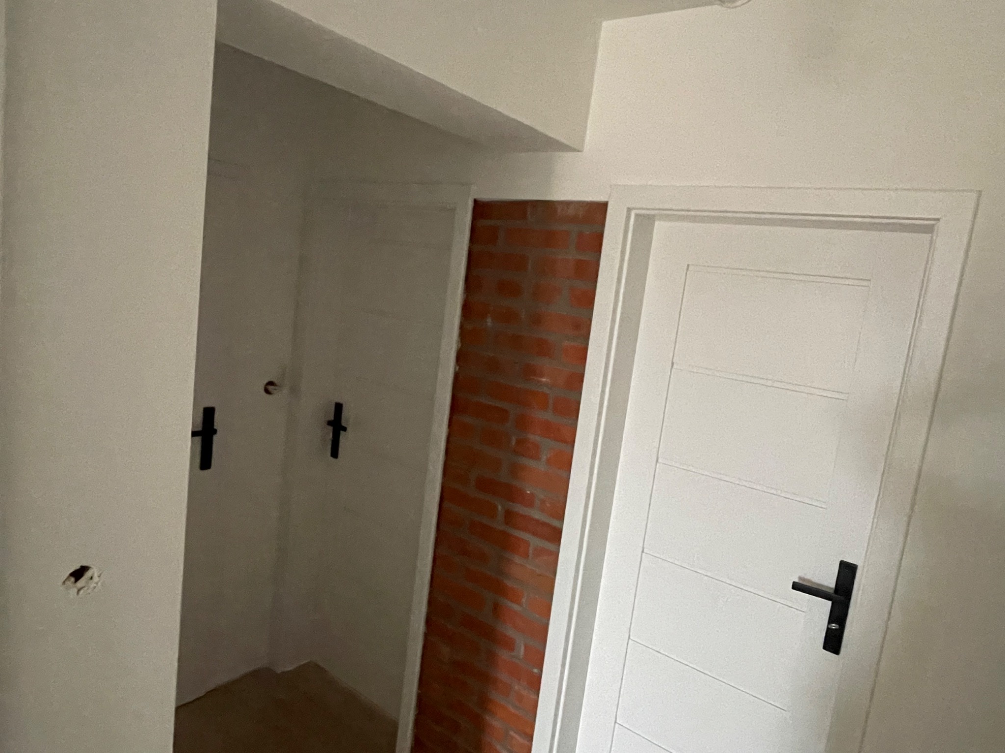 Wnętrze w trakcie remontu: białe ściany, widoczna instalacja elektryczna, dwie białe drzwi z czarnymi klamkami oraz fragment ściany z cegły.