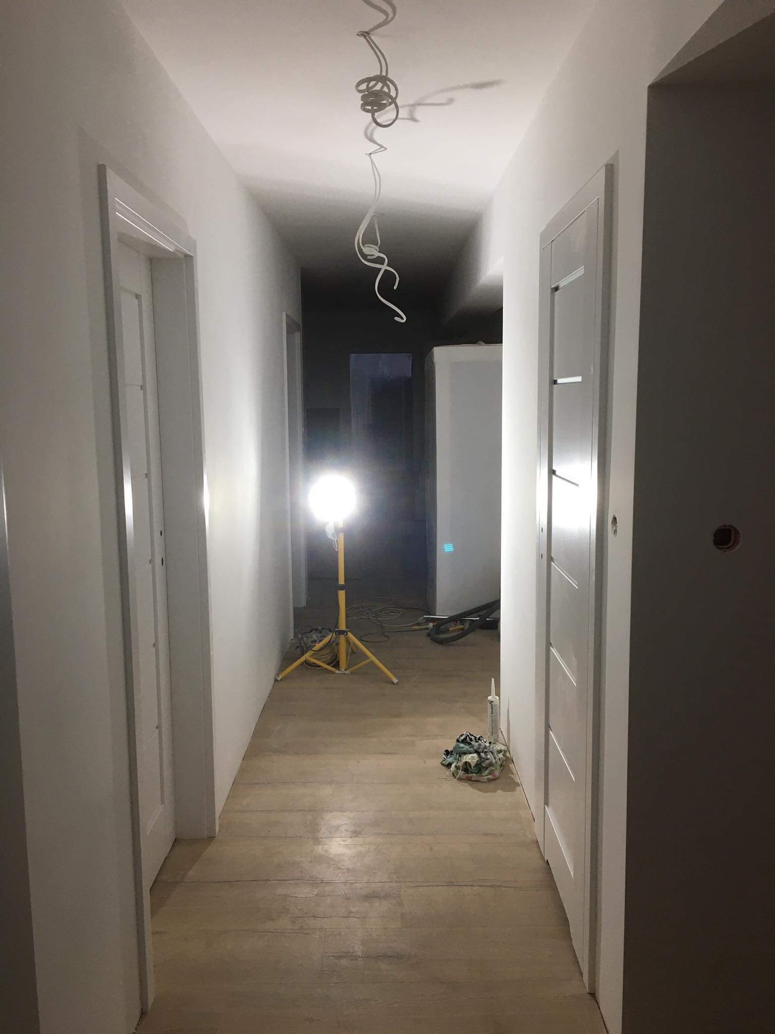 Korytarz w trakcie remontu: świeżo pomalowane białe ściany, drewniana podłoga, zamontowane białe drzwi z poziomymi frezami, żółty halogen na statywie oświetlający przestrzeń, zwisające kable z sufitu.