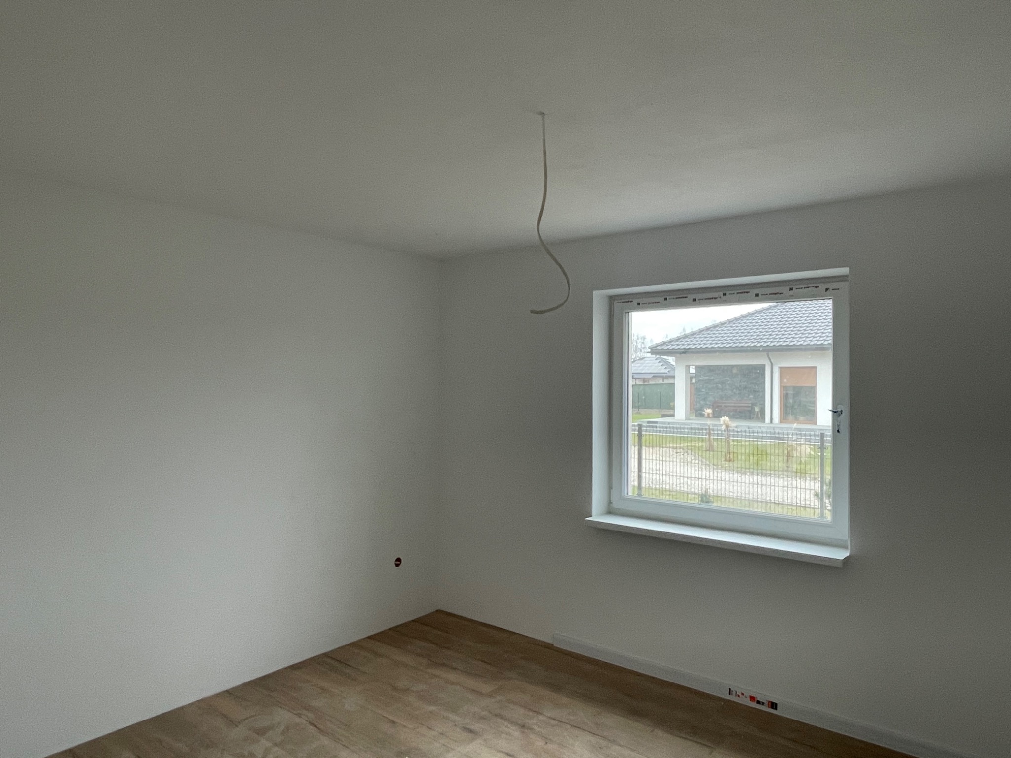 Pusty pokój z drewnopodobną podłogą, świeżo pomalowanymi białymi ścianami i białym oknem z widokiem na dom i ogród. Z sufitu zwisa kabel.