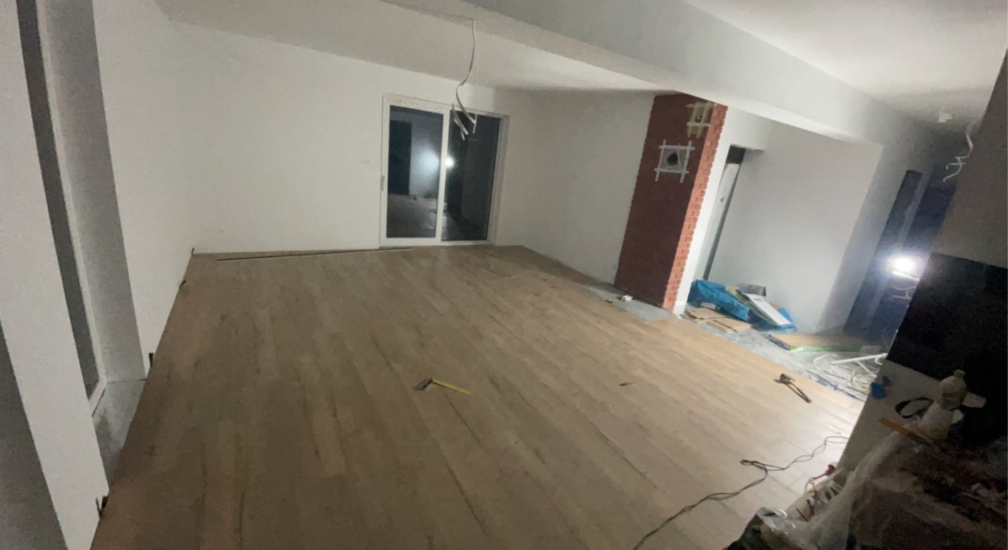 Wnętrze w trakcie prac wykończeniowych: świeżo położona drewniana podłoga, białe ściany, częściowo wykończona ściana z cegły, widoczne narzędzia i materiały budowlane.