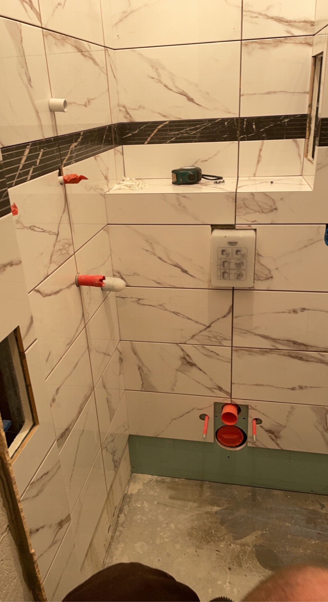 Łazienka w trakcie remontu: białe płytki marmurowe z czarnym pasem, widoczne rury i instalacja podtynkowa, przymocowany stelaż podtynkowy WC, poziomica oraz resztki zaprawy na półce.