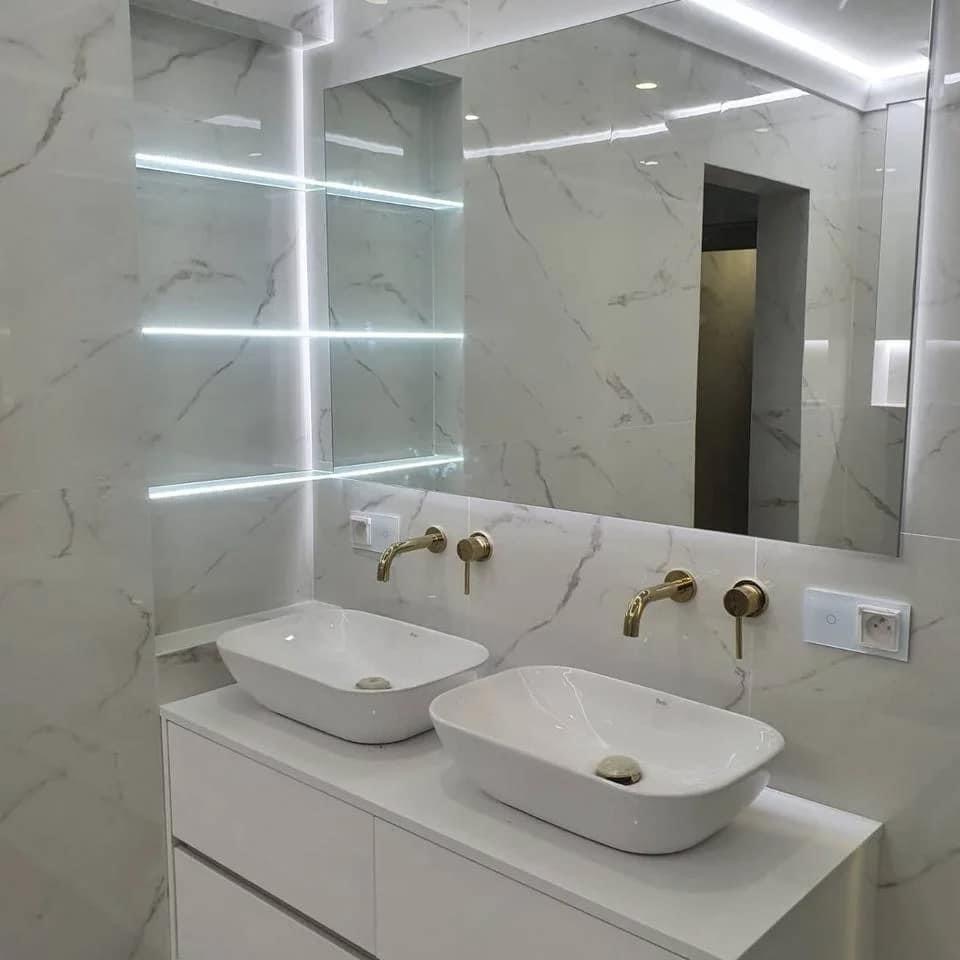 Nowoczesna łazienka z dwoma umywalkami nablatowymi, złotą armaturą i podświetlanym lustrem w marmurowej okładzinie.