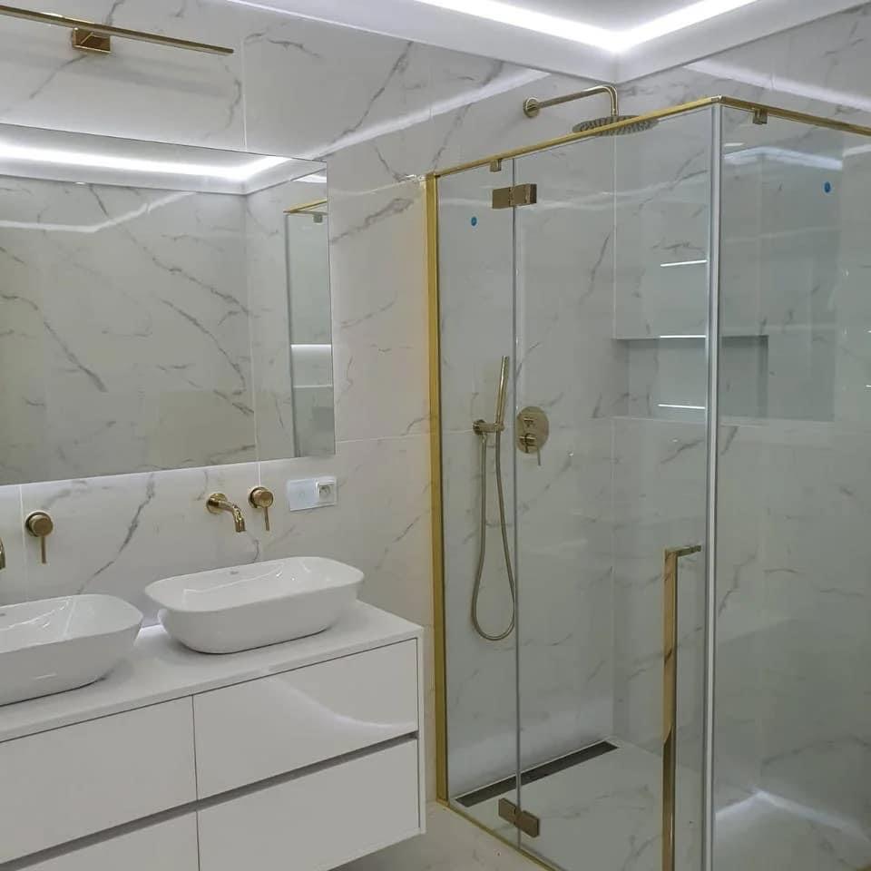 Nowoczesna łazienka z marmurowymi płytkami, złotymi detalami armatury i kabiny prysznicowej, podwójna umywalka nablatowa na białej szafce, oświetlenie LED.