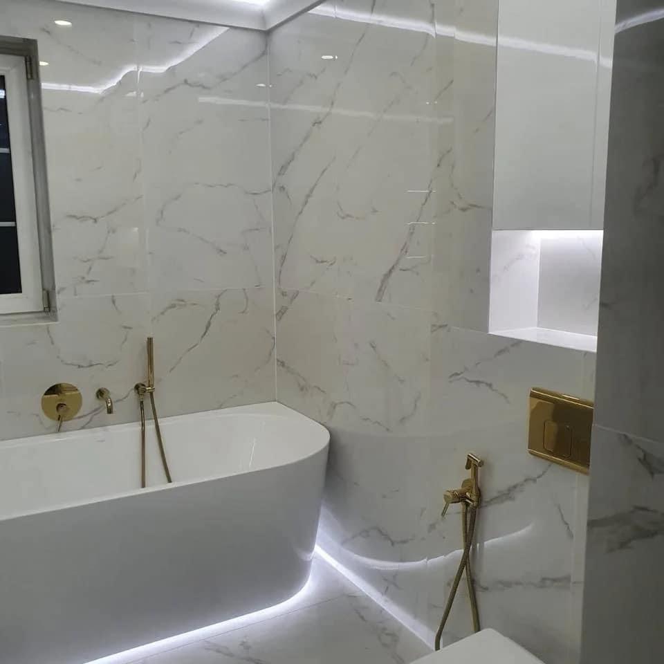 Nowoczesna łazienka z białymi marmurowymi płytkami, podświetlaną wanną wolnostojącą i złotą armaturą, oświetlenie LED pod wanną i w półce ściennej.