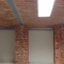 Ceglany sufit z widocznymi belkami wzmacniającymi i prostokątnym oknem dachowym, wsparty na dwóch ceglanych kolumnach na tle gładkiej, pomalowanej ściany.