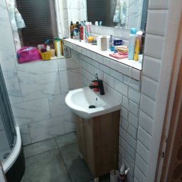 Perfectfinish - Umywalka z szafką w łazience wyłożonej białymi, prostokątnymi płytkami. W tle kabina prysznicowa i okno z żaluzjami. Na półce nad umywalką kosmetyki i akcesoria.