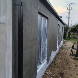 Perfectfinish - Ściana budynku w trakcie prac wykończeniowych: świeżo nałożony tynk, okna zabezpieczone folią, widoczny system rynnowy. Ujęcie z perspektywy ziemi.