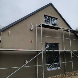 Perfectfinish - Elewacja domu w trakcie prac wykończeniowych: świeżo nałożony tynk w odcieniach szarości, okna zabezpieczone folią, widoczne rusztowania. Detale instalacji na ścianie.