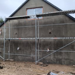 Perfectfinish - Świeżo otynkowana ściana domu jednorodzinnego z widocznymi rusztowaniami i zabezpieczonym oknem, ukazująca etap prac wykończeniowych na zewnątrz budynku.