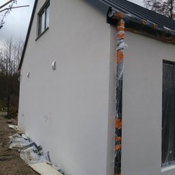 Perfectfinish - Elewacja budynku po nałożeniu tynku strukturalnego, z zabezpieczonymi elementami (rynna, okna) folią i taśmą. Widoczne fragmenty instalacji i resztki materiałów budowlanych.