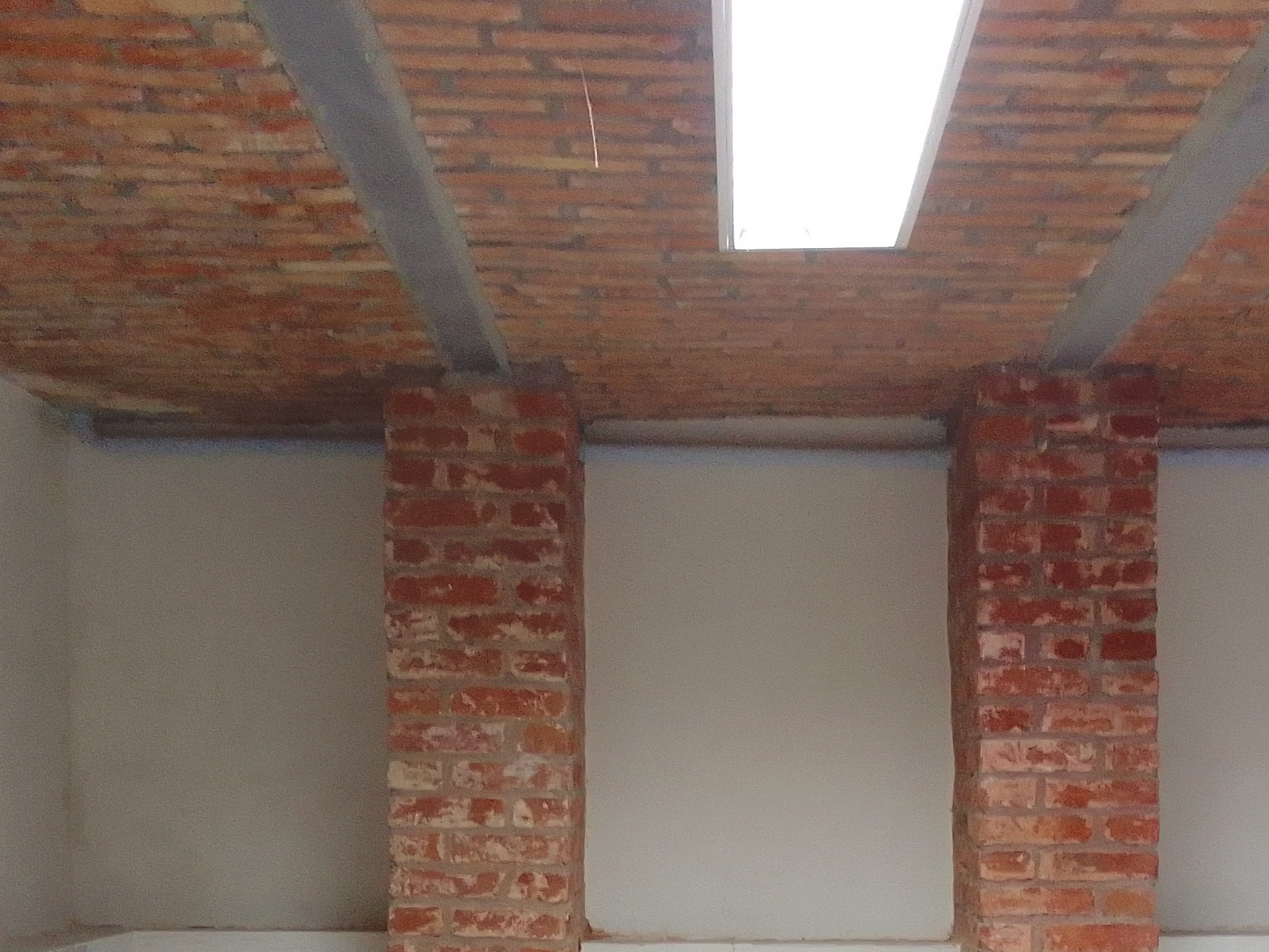 Ceglany sufit z widocznymi belkami wzmacniającymi i prostokątnym oknem dachowym, wsparty na dwóch ceglanych kolumnach na tle gładkiej, pomalowanej ściany.