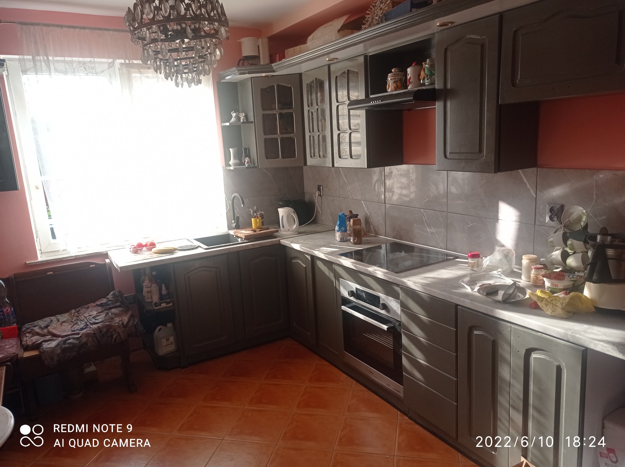 Wykończona kuchnia z szarymi szafkami, marmurowymi płytkami na ścianie i brązowymi płytkami na podłodze, z widocznym oknem i ozdobnym żyrandolem.