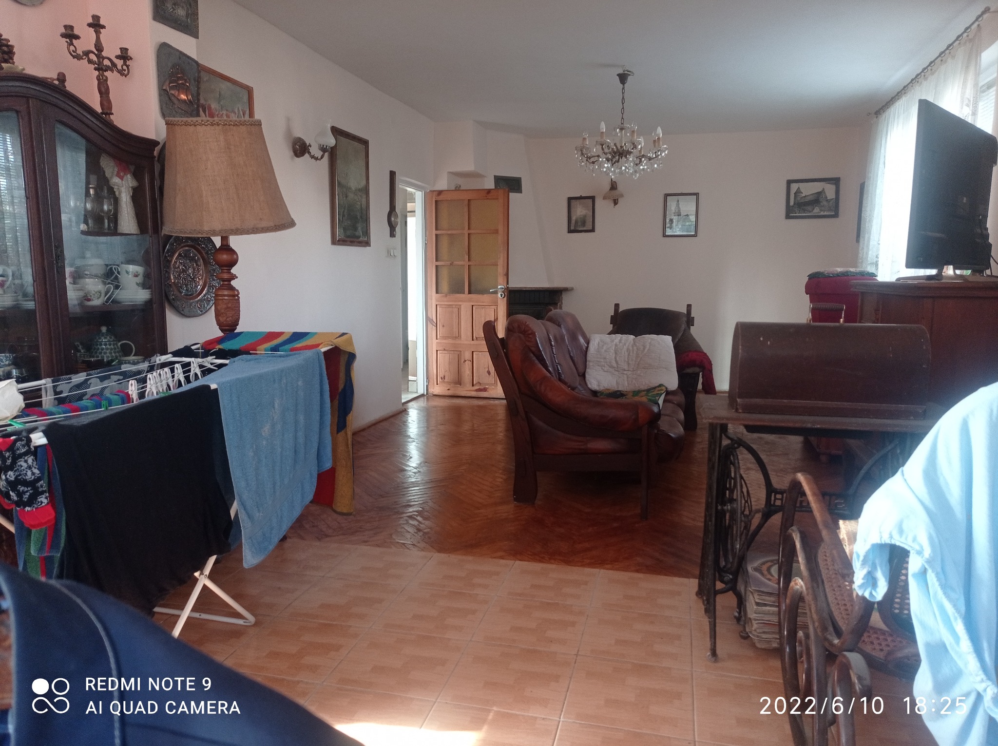 Widok na przestronne wnętrze pokoju dziennego z brązowymi płytkami podłogowymi, staromodnymi meblami, w tym skórzaną sofą i kredensem, oraz suszarką na pranie z kolorowymi ubraniami.