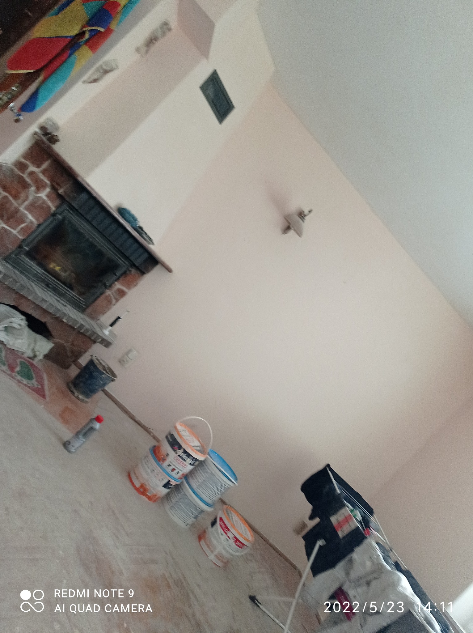 Wnętrze w trakcie malowania ścian na jasny kolor, widoczne puszki z farbą na podłodze, odsłonięty kominek z cegły i lampa sufitowa, ujęcie z dołu.