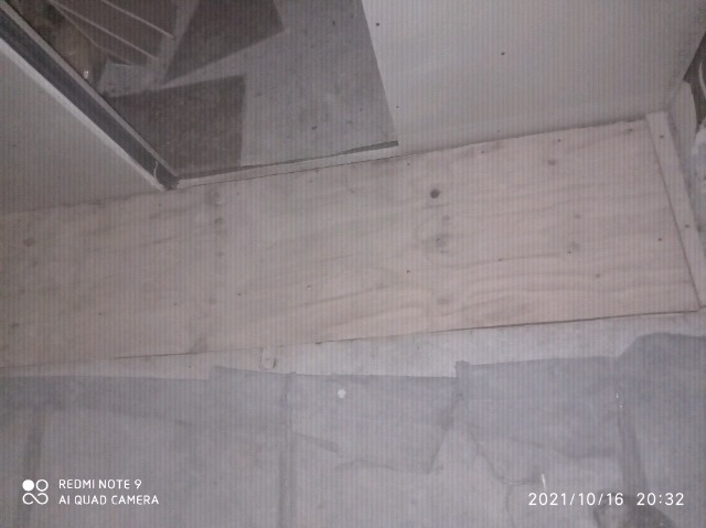 Surowe deski tworzące podest pod schodami, widoczne fragmenty betonowej podłogi i schodów, zdjęcie wykonane z góry, słabe oświetlenie.