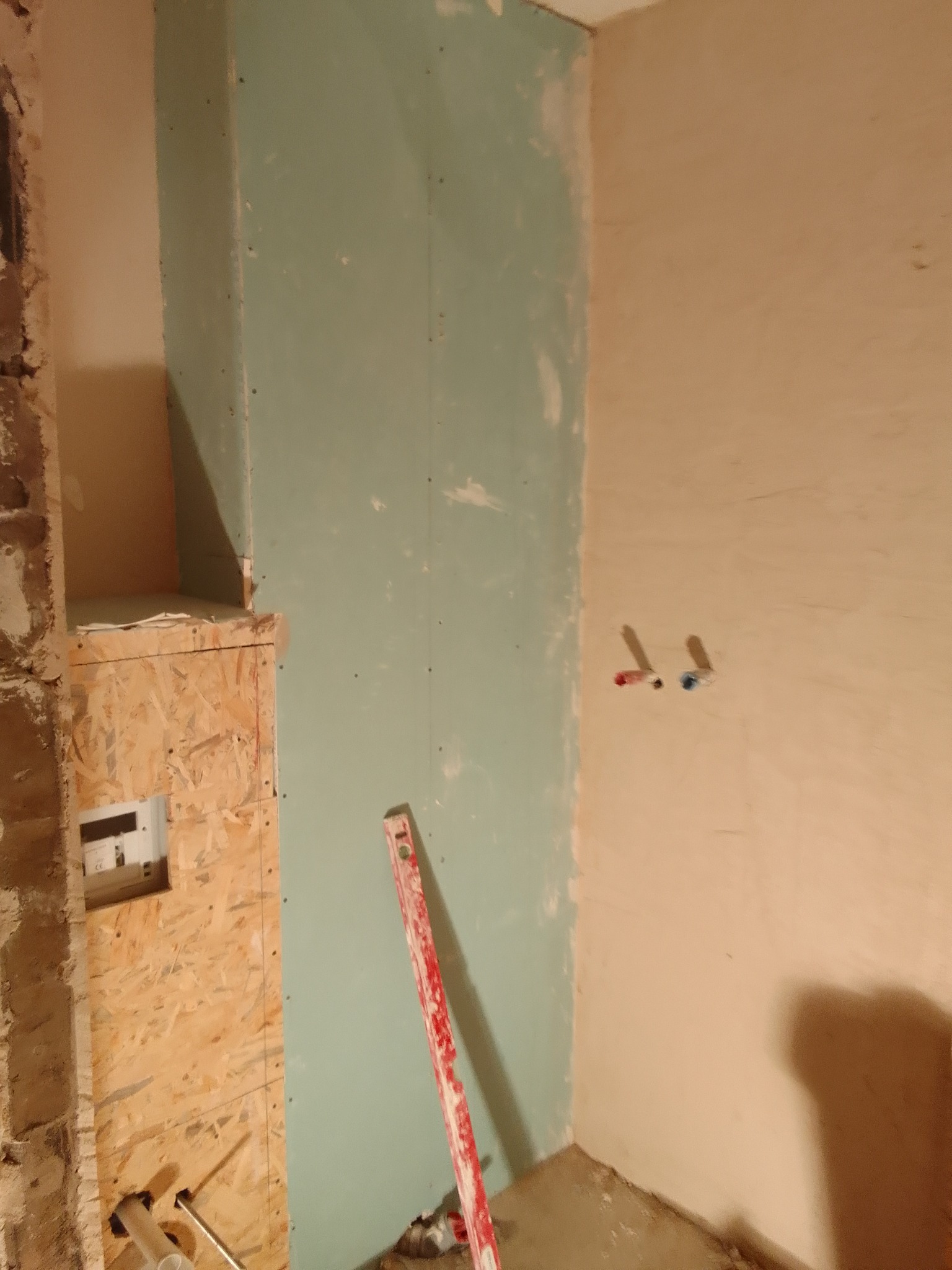 Niedokończona łazienka w trakcie remontu: widoczne płyty gipsowo-kartonowe na ścianach, pionowa poziomica oparta o ścianę i fragment instalacji wodnej z wystającymi rurkami.