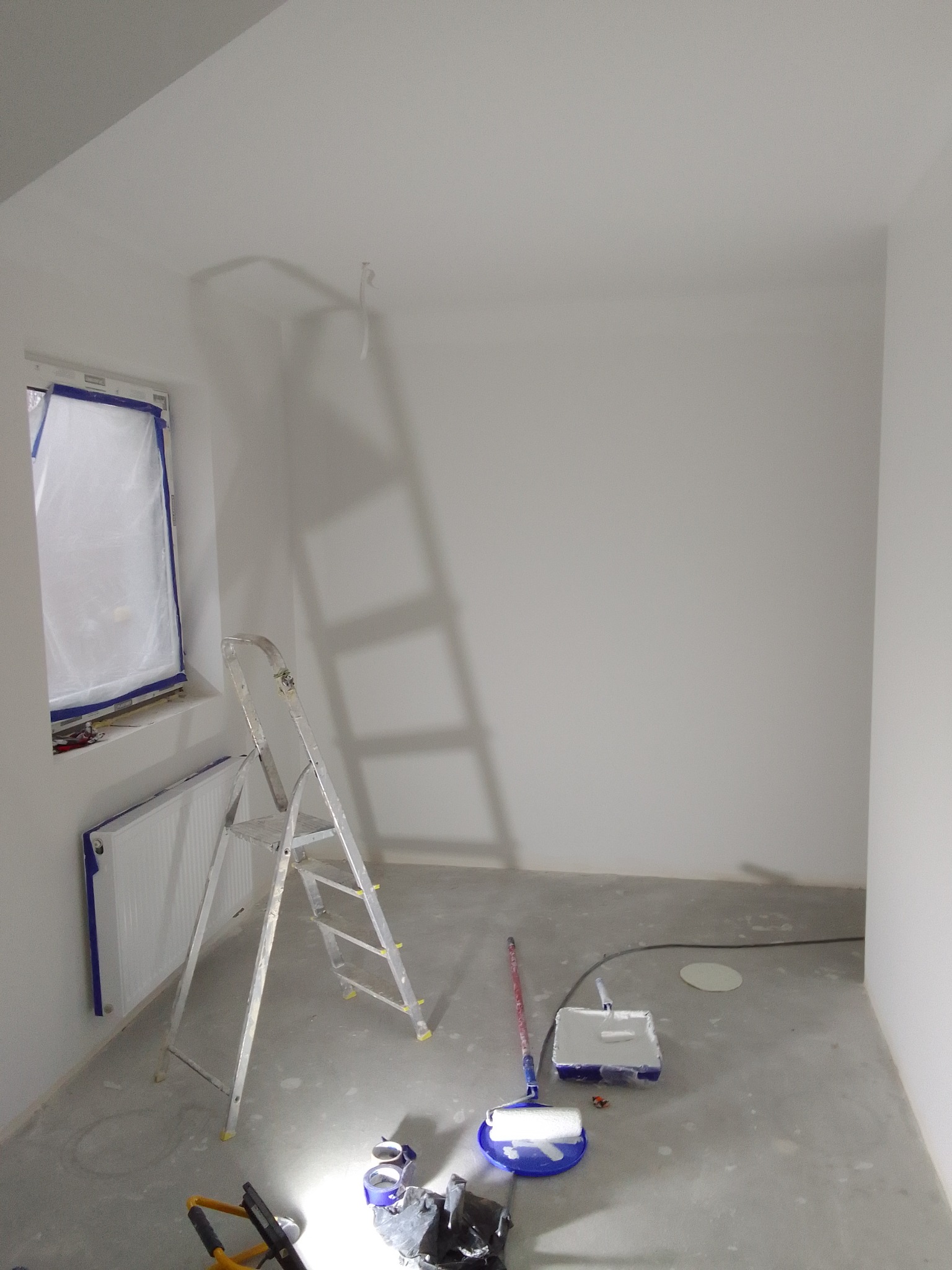 Pokój w trakcie malowania na biało, okno i grzejnik zabezpieczone folią, drabina aluminiowa, kuweta z wałkiem malarskim i inne narzędzia na podłodze.