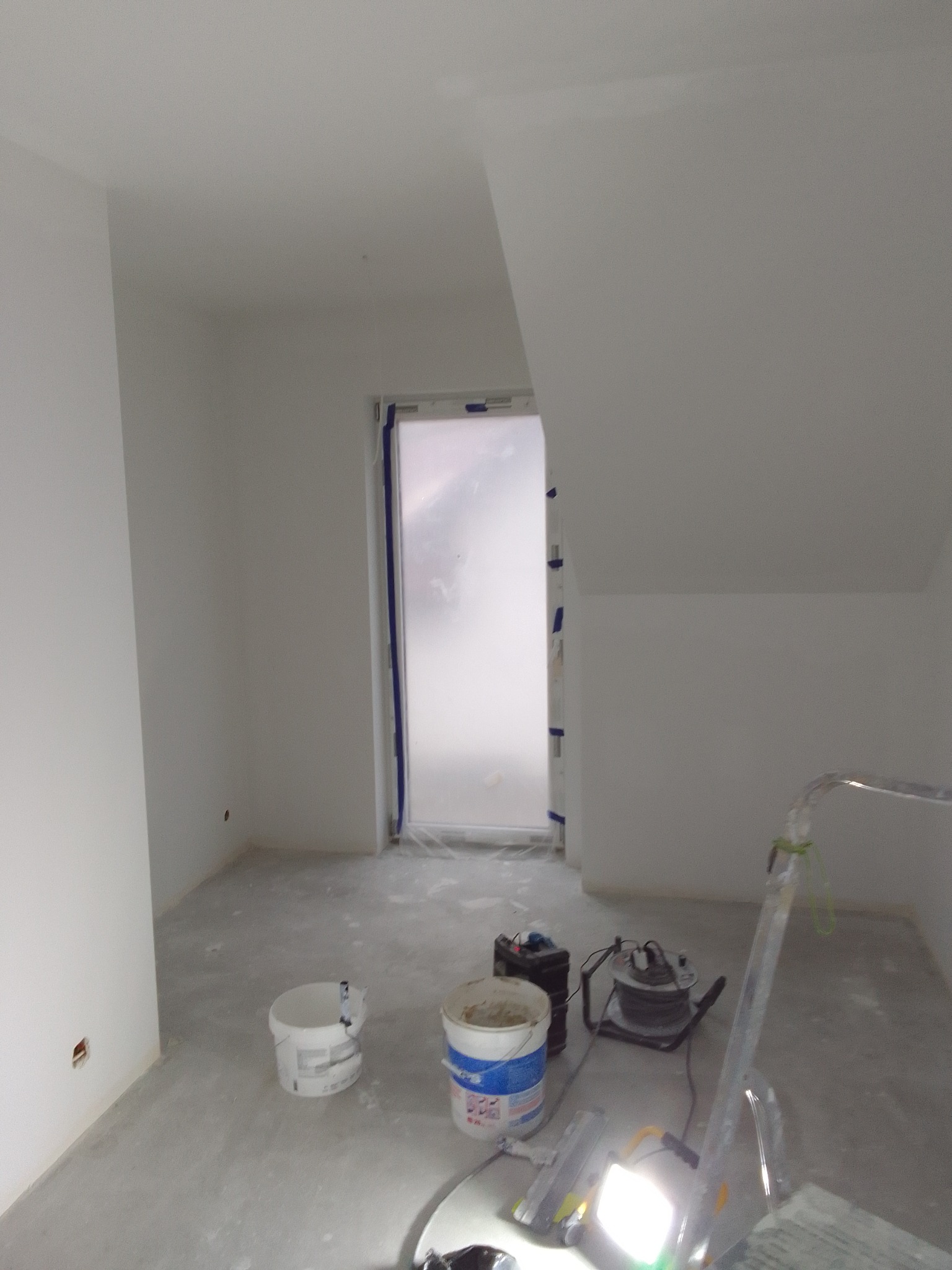 Pokój w trakcie malowania: świeżo pomalowane na biało ściany i sufit, okno zabezpieczone folią malarską, widoczne dwa wiadra z farbą, szlifierka do gładzi i lampa budowlana na statywie.