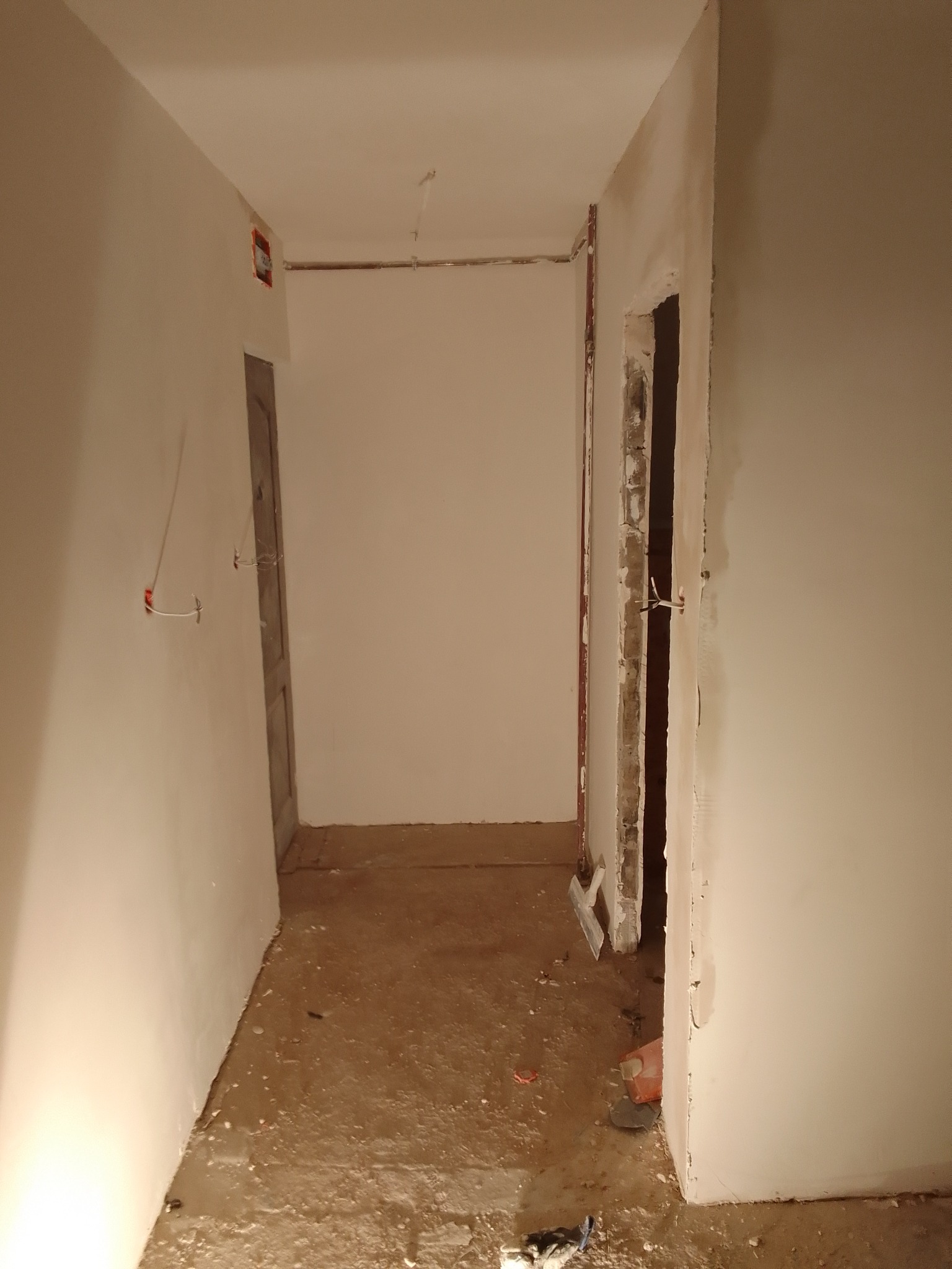 Korytarz w trakcie prac remontowych w Olsztynie: surowe ściany po skrobaniu, widoczne instalacje elektryczne, podłoga przygotowana do dalszych prac wykończeniowych, szpachelka oparta o ścianę.