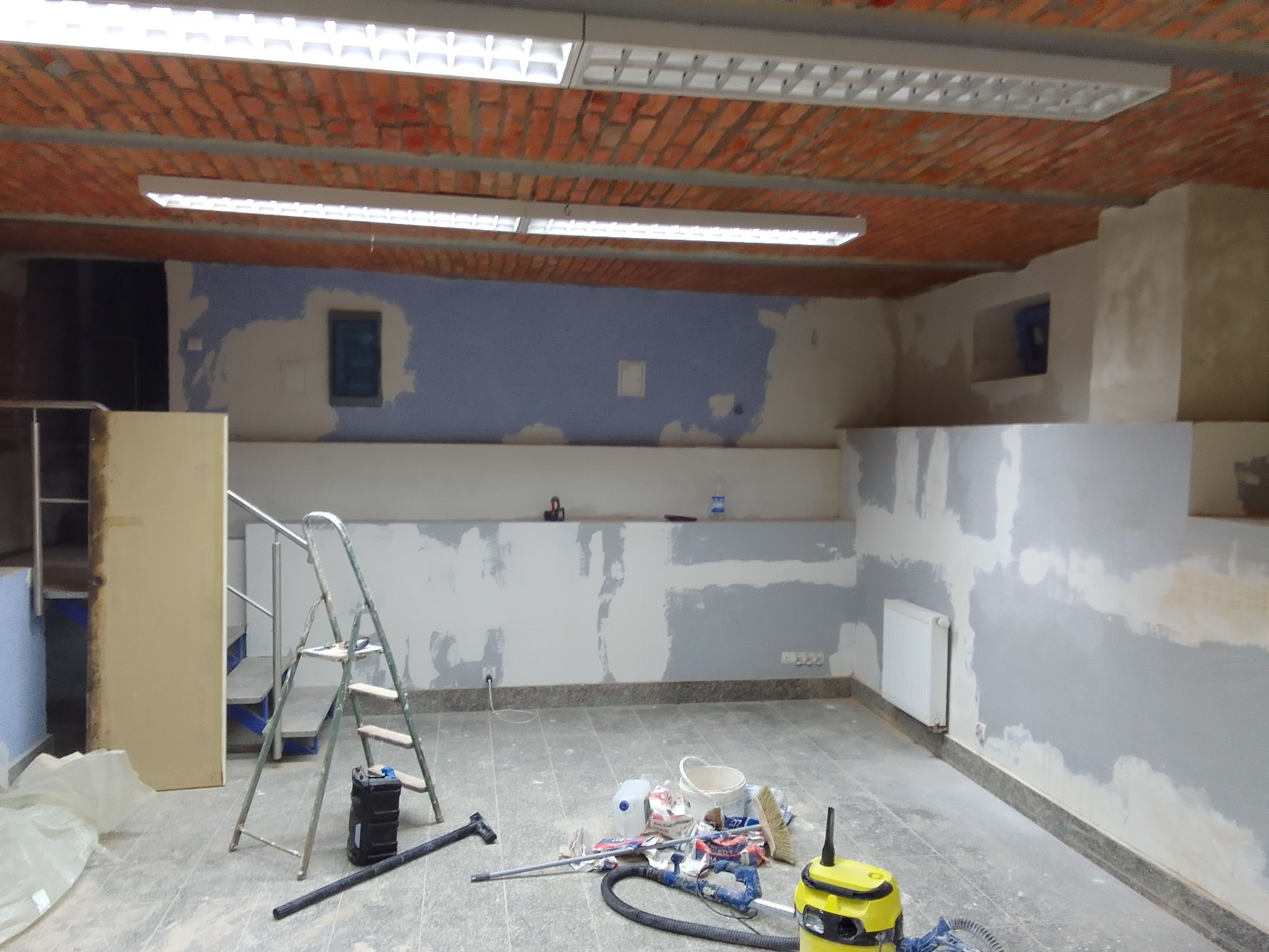 Pomieszczenie biurowe w trakcie przygotowania do malowania: ściany częściowo zagruntowane, widoczna drabina, narzędzia malarskie i odkurzacz przemysłowy na podłodze, surowy ceglany sufit...