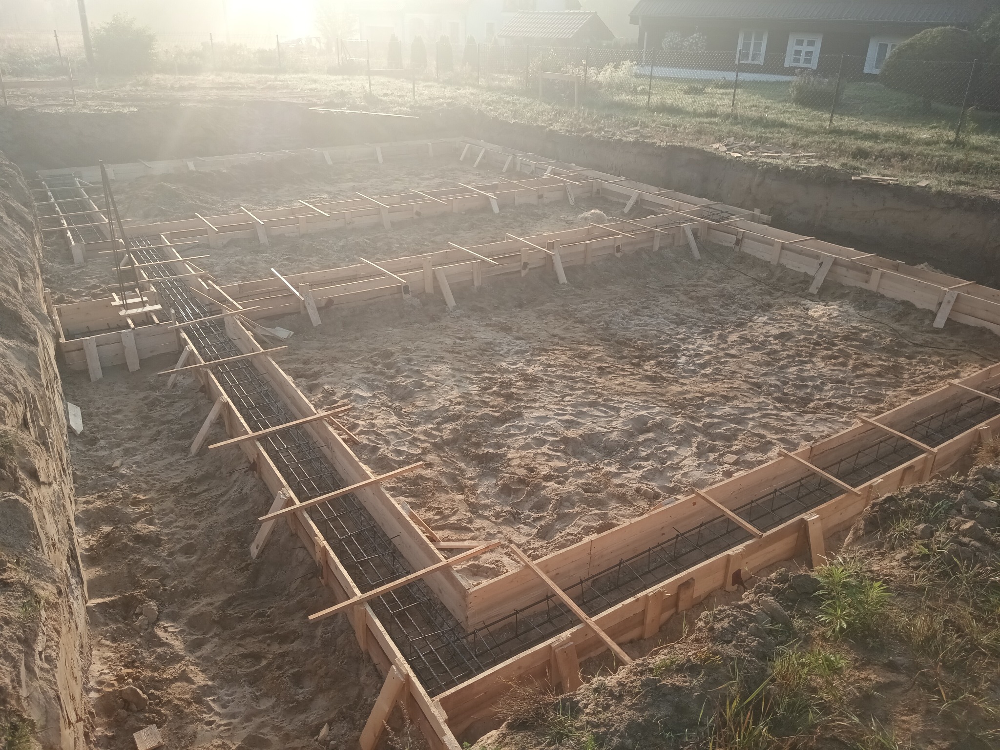Przygotowanie fundamentów pod budynek: drewniane szalunki zbrojone stalą, wypełnione piaskiem, w wykopie ziemnym, w porannym świetle. Widok z góry.