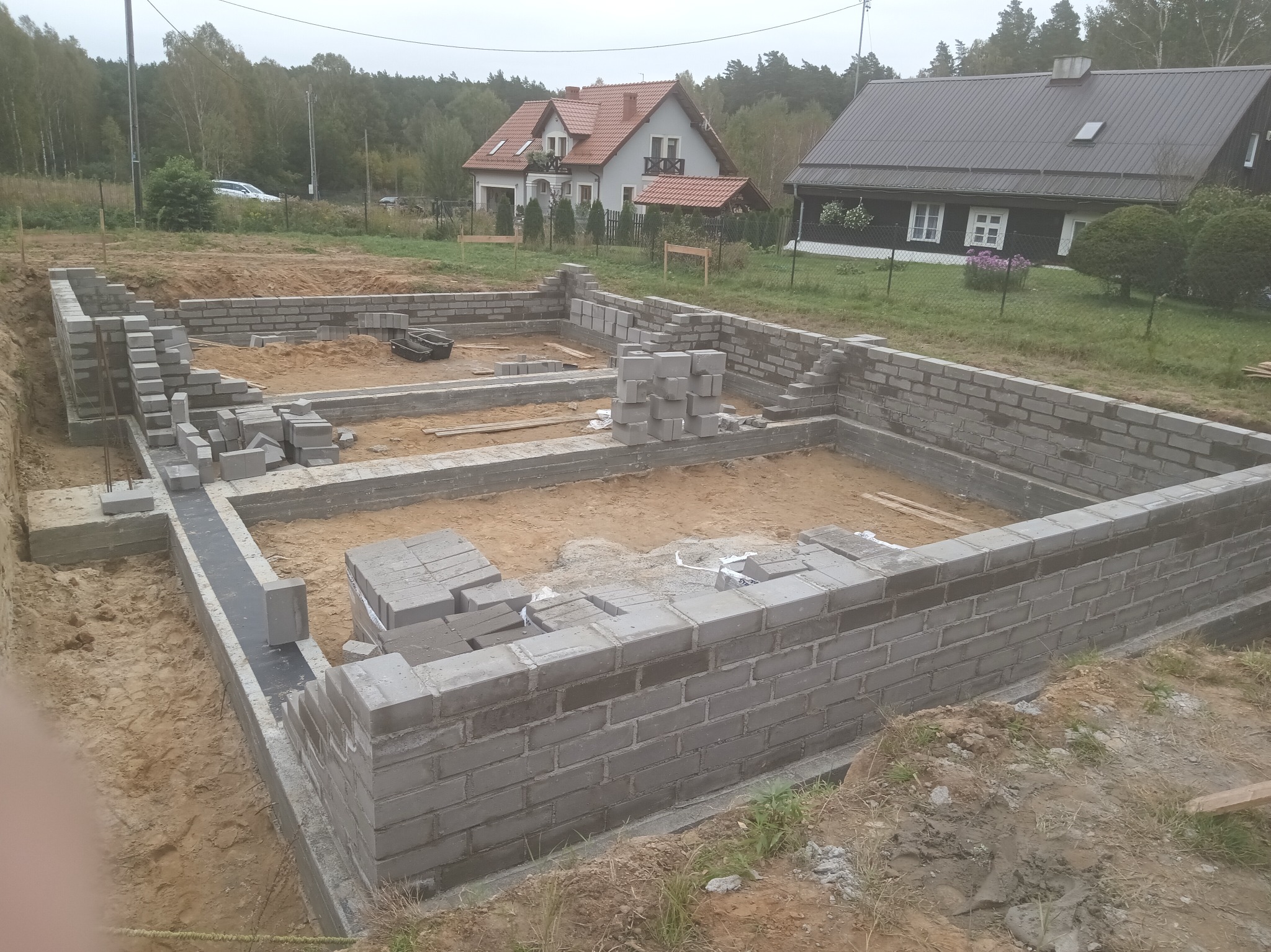 Fundamenty domu w trakcie budowy, widoczne bloczki betonowe i izolacja, w tle dwa domy jednorodzinne i drzewa. Ujęcie z góry, częściowo widoczna dłoń w lewym dolnym rogu.